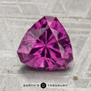 A 2.30-carat pastel pyrope garnet, trillion-cut and purplish-pink, rests on a textured gray surface with "Earth's Treasury" shown at the bottom.