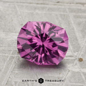 A 2.21-carat pastel pyrope garnet, faceted in a round cut, rests on a textured gray surface with "EARTH'S TREASURY" displayed below.