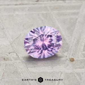 A round brilliant-cut 0.78-carat Montana Sapphire with a purple hue is displayed on a textured metallic surface. The "EARTH'S TREASURY" logo is shown at the bottom.