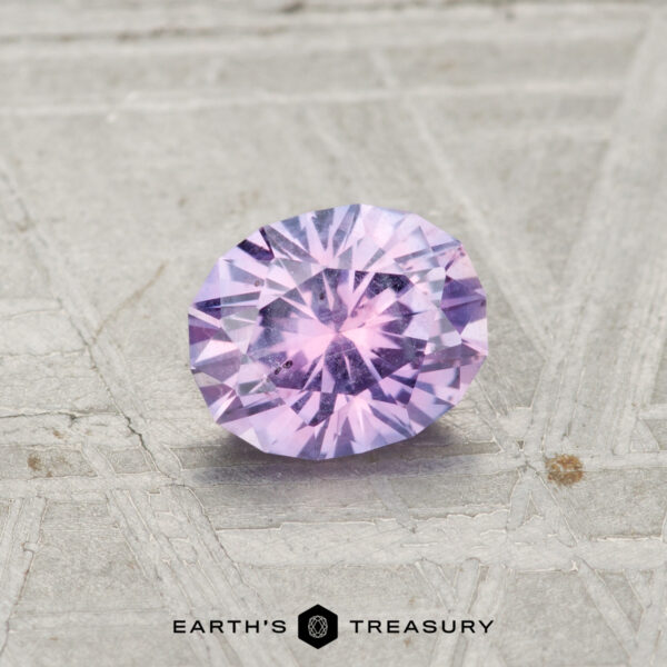0.78-Carat Pinkish Purple Montana Sapphire A round brilliant-cut 0.78-carat Montana Sapphire with a purple hue is displayed on a textured metallic surface. The "EARTH'S TREASURY" logo is shown at the bottom.