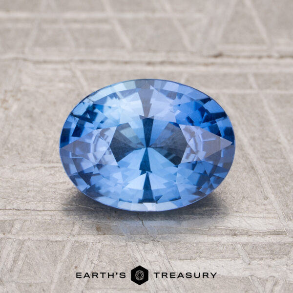 1.77-Carat Cornflower Blue Ceylon Sapphire A 1.77-carat Ceylon sapphire, oval-cut and blue, rests on a textured light gray surface with the "Earth's Treasury" logo and text displayed at the bottom of the image.