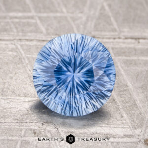 A 1.85-carat unheated Ceylon sapphire, round brilliant-cut, is showcased on a textured light surface with the "Earth's Treasury" logo and text below.