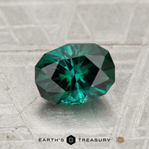 A 1.23-carat faceted green tourmaline with a starburst pattern rests on a textured gray surface, with "Earth's Treasury" written below.