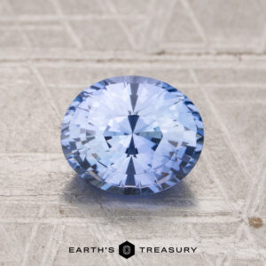 A round, faceted 1.69-carat Ceylon sapphire with a light blue hue is displayed on a textured beige surface with the "Earth's Treasury" logo below.