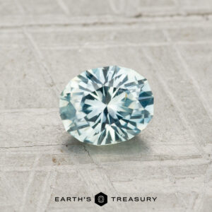 A 0.77-carat, round, faceted pale blue Montana sapphire is displayed on a textured gray surface with the text "Earth's Treasury" at the bottom.
