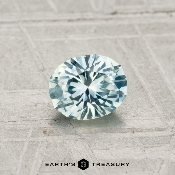 0.77-Carat Light Aqua Montana Sapphire A 0.77-carat, round, faceted pale blue Montana sapphire is displayed on a textured gray surface with the text "Earth's Treasury" at the bottom.