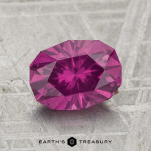 A 2.16-carat pastel pyrope garnet, faceted in an oval shape, rests on a textured metallic surface featuring the "EARTH'S TREASURY" text and logo below.