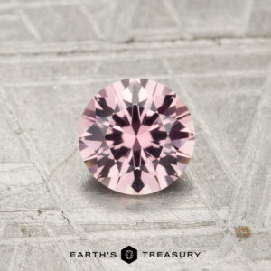 A 0.74-carat round-cut Montana sapphire shines on a textured gray surface with the "Earth's Treasury" logo at the bottom.