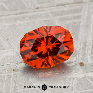 A 2.08-carat Spessartine Garnet, showcasing a vivid orange-red hue and faceted cut, rests on a textured gray surface with "EARTH'S TREASURY" displayed at the bottom.