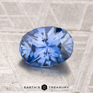 A 1.67-carat heated Ceylon sapphire is displayed on a textured metallic surface, with the text "EARTH'S TREASURY" appearing at the bottom of the image.