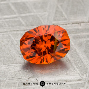 A 1.86-carat faceted spessartine garnet with a brilliant cut is displayed on a textured gray surface, with the "Earth's Treasury" logo at the bottom.