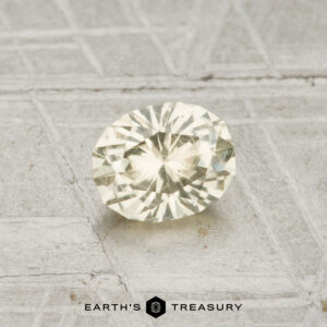 A 0.74-carat Montana sapphire with an oval cut and yellow hue is displayed on a textured gray surface, featuring the text "EARTH'S TREASURY" at the bottom.