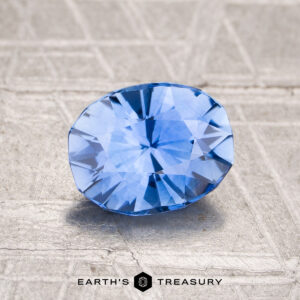 A 1.59-carat heated Ceylon sapphire, cut in a faceted oval, rests on a textured light surface with "EARTH'S TREASURY" at the bottom.