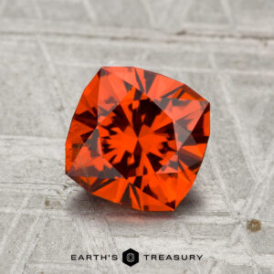 A 1.74-carat Spessartine garnet with vivid orange facets sits on a textured gray surface, featuring the "Earth's Treasury" name and hexagon logo at the bottom.