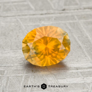A 1.40-carat Montana Sapphire, faceted in an oval cut with a yellow-orange hue, is displayed on a textured light gray surface with the Earth’s Treasury text and logo at the bottom.
