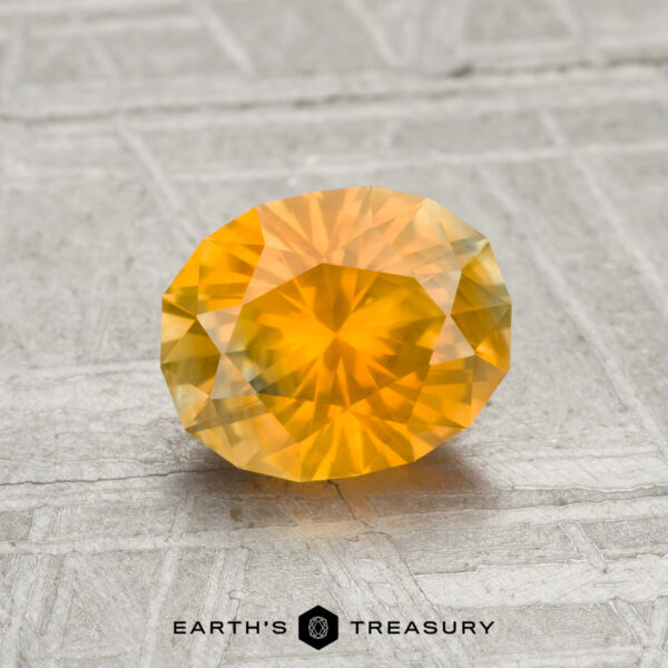 1.40-Carat Orange-Yellow Particolored Montana Sapphire A 1.40-carat Montana Sapphire, faceted in an oval cut with a yellow-orange hue, is displayed on a textured light gray surface with the Earth’s Treasury text and logo at the bottom.