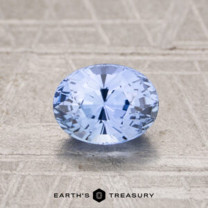 A 1.24-carat Ceylon sapphire, cut in a faceted oval shape and light blue in color, rests on a textured beige surface with "EARTH'S TREASURY" displayed at the bottom.