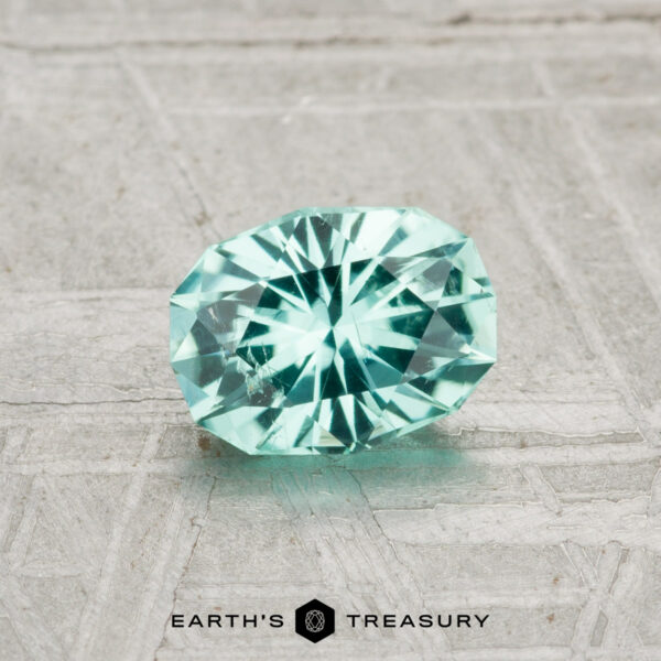 0.79-Carat Tourmaline A faceted, oval-cut 0.79-carat light blue tourmaline rests on a textured gray surface, with the "Earth's Treasury" logo displayed at the bottom.