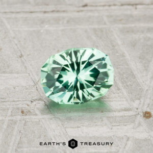A 0.59-carat faceted oval green tourmaline rests on a textured gray surface, with the Earth's Treasury logo at the bottom.