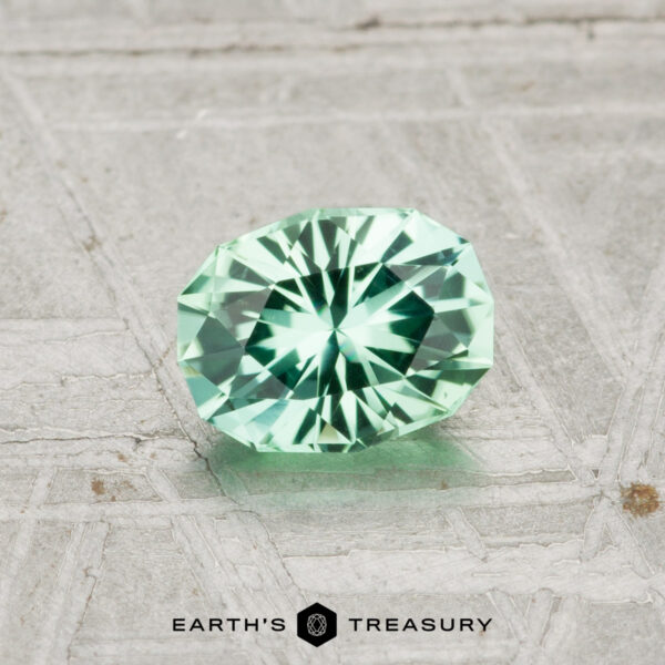 0.59-Carat Tourmaline A 0.59-carat faceted oval green tourmaline rests on a textured gray surface, with the Earth's Treasury logo at the bottom.