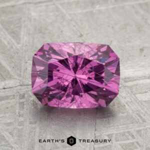A rectangular, faceted 2.01-carat pastel pyrope garnet with visible inclusions sits on a gray patterned background, with "EARTH'S TREASURY" beneath it.