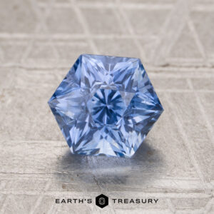 A 2.06-carat Ceylon sapphire with hexagonal facets is displayed on a textured, light surface, with "Earth's Treasury" written below.