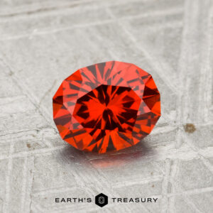 A 1.28-carat Spessartine garnet, faceted in an oval shape, is displayed on a textured gray surface with the Earth’s Treasury logo below.