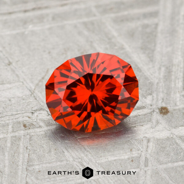 1.28-Carat Spessartine Garnet A 1.28-carat Spessartine garnet, faceted in an oval shape, is displayed on a textured gray surface with the Earth’s Treasury logo below.