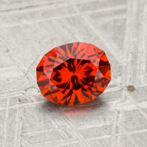 A 1.28-carat Spessartine garnet, faceted in an oval shape, is displayed on a textured gray surface with the Earth’s Treasury logo below.