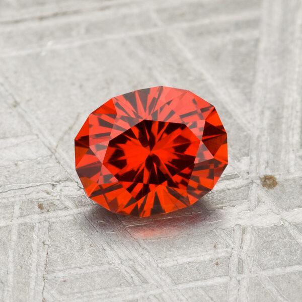 A 1.28-carat Spessartine garnet, faceted in an oval shape, is displayed on a textured gray surface with the Earth’s Treasury logo below.