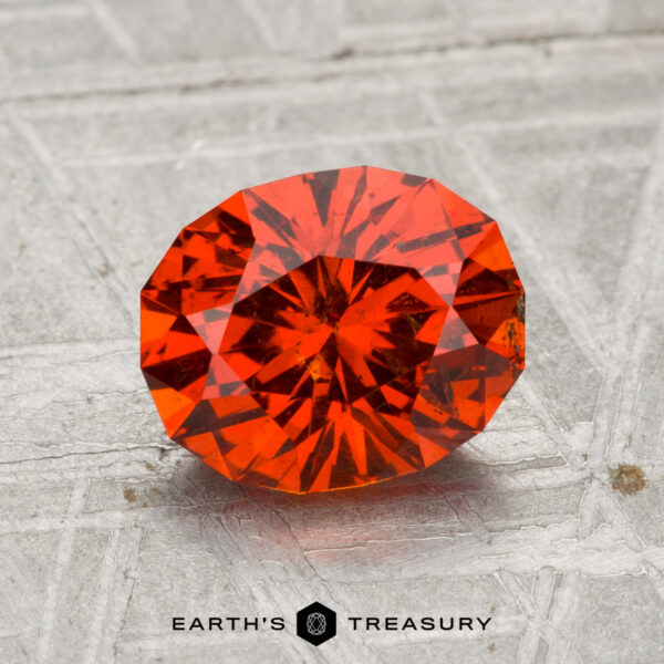 2.11-Carat Spessartine Garnet A 2.11-carat spessartine garnet, a faceted oval orange-red gemstone, rests on a textured gray surface with the text "EARTH'S TREASURY" below.