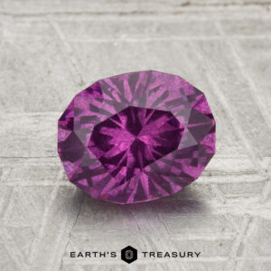 A 1.92-carat pastel pyrope garnet with an oval cut is displayed on a gray textured surface, featuring the "EARTH'S TREASURY" text at the bottom.