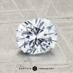 A faceted, oval-shaped 1.89-carat Montana sapphire with a clear, reflective surface rests on a textured gray background. "Earth's Treasury" appears below the stone.