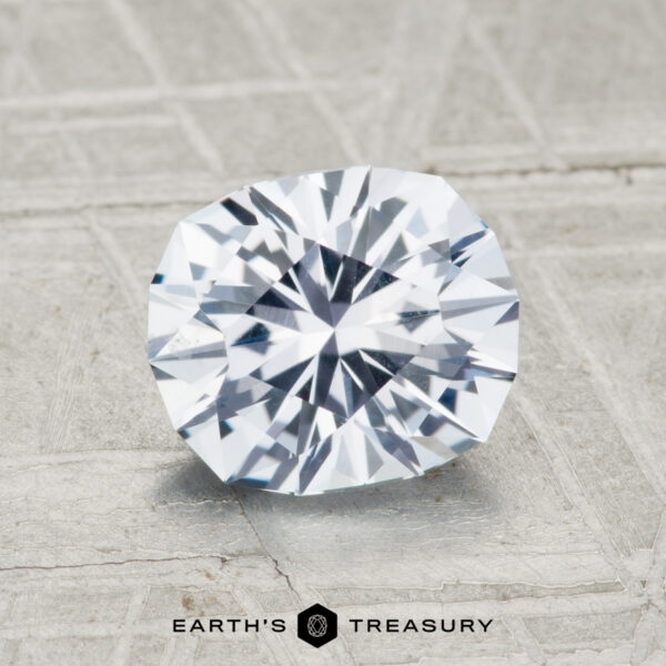 1.89-Carat Near-Colorless Montana Sapphire A faceted, oval-shaped 1.89-carat Montana sapphire with a clear, reflective surface rests on a textured gray background. "Earth's Treasury" appears below the stone.