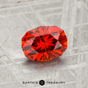 A faceted, oval-shaped 1.31-carat Spessartine Garnet displayed on a textured gray surface with "EARTH'S TREASURY" text at the bottom.