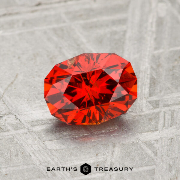1.31-Carat Spessartine Garnet A faceted, oval-shaped 1.31-carat Spessartine Garnet displayed on a textured gray surface with "EARTH'S TREASURY" text at the bottom.