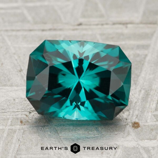2.44-Carat Indicolite Tourmaline A 2.44-carat Indicolite Tourmaline with an emerald cut is displayed on a textured gray surface. The teal-colored, faceted gemstone sits above the "EARTH'S TREASURY" logo at the bottom of the image.
