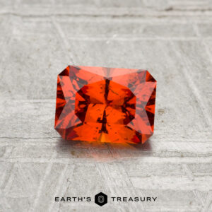 A 0.97-carat rectangular, faceted Spessartine Garnet with an orange hue is shown on a textured gray surface, featuring the "EARTH'S TREASURY" text and hexagon logo below.