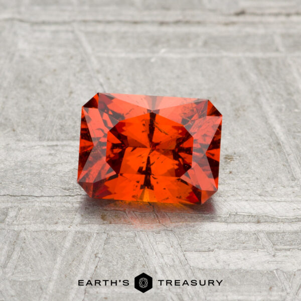0.97-Carat Spessartine Garnet A 0.97-carat rectangular, faceted Spessartine Garnet with an orange hue is shown on a textured gray surface, featuring the "EARTH'S TREASURY" text and hexagon logo below.