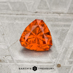 A faceted, triangular 0.85-carat Spessartine Garnet rests on a textured metallic surface with "EARTH'S TREASURY" and a hexagon logo at the bottom.