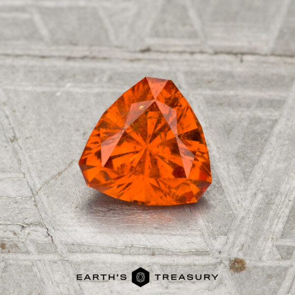 0.85-Carat Spessartine Garnet A faceted, triangular 0.85-carat Spessartine Garnet rests on a textured metallic surface with "EARTH'S TREASURY" and a hexagon logo at the bottom.