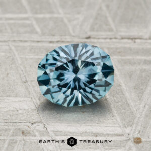 A 1.21-carat Montana Sapphire, oval-cut with radiant facets, rests on a textured, light background. "EARTH'S TREASURY" is displayed at the bottom of the image.