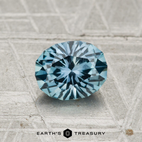 1.21-Carat Teal Blue Montana Sapphire A 1.21-carat Montana Sapphire, oval-cut with radiant facets, rests on a textured, light background. "EARTH'S TREASURY" is displayed at the bottom of the image.