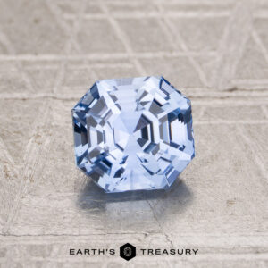 The 1.55-Carat Ceylon Sapphire, a faceted octagonal blue gemstone, is displayed on a textured metallic surface with "EARTH'S TREASURY" at the bottom.