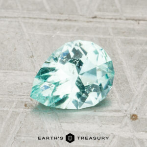 A 1.10-carat Maine tourmaline, pear-shaped and light blue-green, is displayed on a textured gray surface with the text "EARTH'S TREASURY" at the bottom.