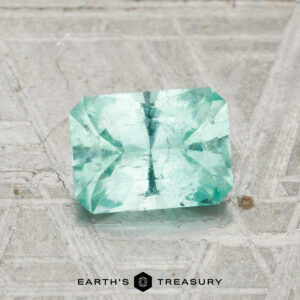 A 1.06-carat Maine tourmaline, rectangular and faceted in teal, rests on a textured gray surface with geometric patterns; "EARTH'S TREASURY" logo appears below.