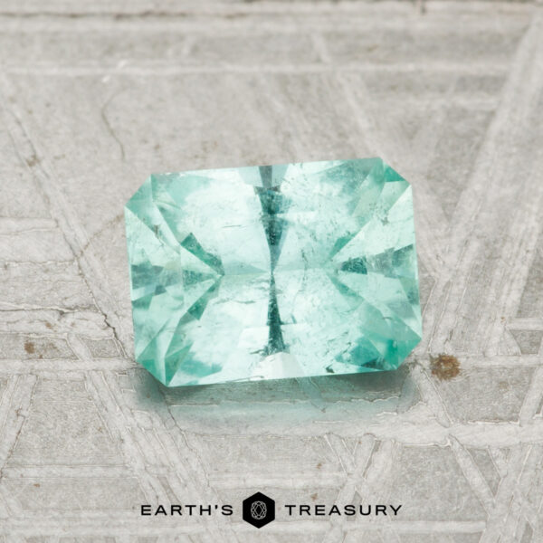 1.06-Carat Maine Tourmaline A 1.06-carat Maine tourmaline, rectangular and faceted in teal, rests on a textured gray surface with geometric patterns; "EARTH'S TREASURY" logo appears below.