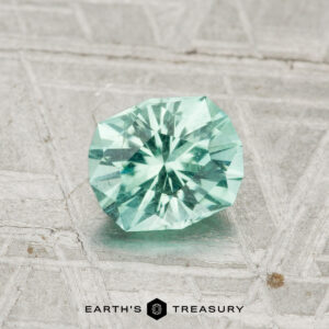 A round, faceted 0.85-carat Maine Tourmaline in light teal is displayed on a textured, silvery background with the "Earth's Treasury" logo at the bottom.