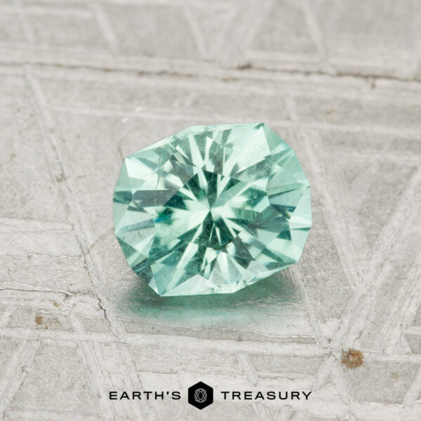 0.85-Carat Maine Tourmaline A round, faceted 0.85-carat Maine Tourmaline in light teal is displayed on a textured, silvery background with the "Earth's Treasury" logo at the bottom.