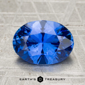 A 3.07-carat heated Ceylon sapphire, oval-cut and blue, displayed on a textured surface with "Earth's Treasury" written below.